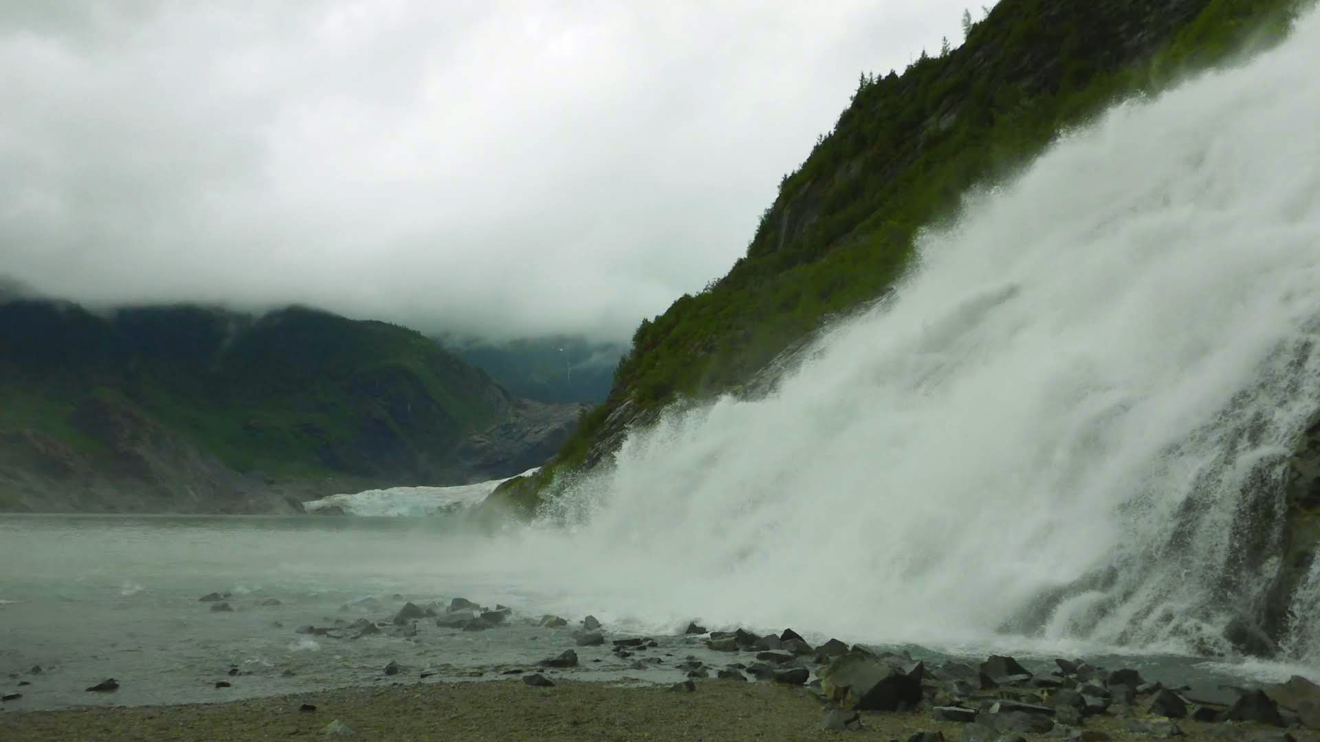 A waterfall tumbles from above against rocks into a glacier fed lake. There is a glacier in the background. It is a dark, overcast and rainy day on an Alaska itinerary