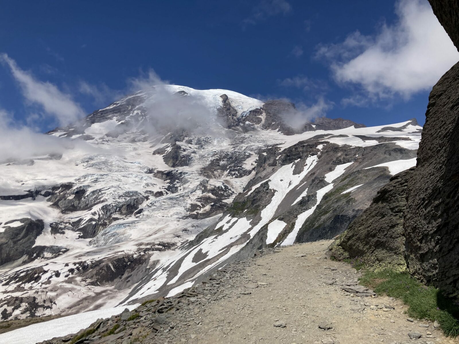 Tahoma or Mt Rainier seen from the Skyline Trail. There is a trail in the foreground and the ice and snow covered mountain rises behind with a few wispy clouds around it on an otherwise clear day. Mt Rainier is a great addition to any seattle itinerary