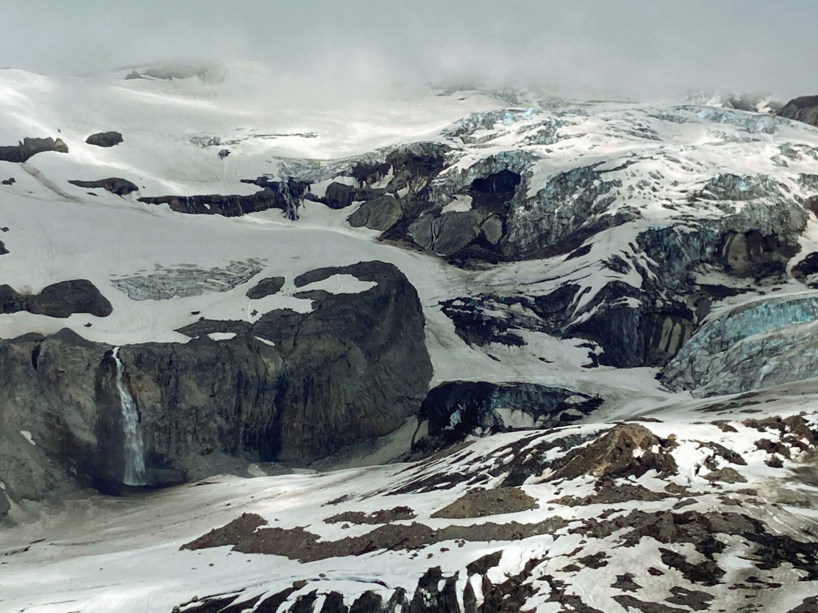 The bottom part of Tahoma/Mt Rainier is visible from the Skyline Trail at Glacier Vista. Below the clouds rocks, snow and glaciers surround the mountain. A high waterfall is visible falling down a rock below a glacier