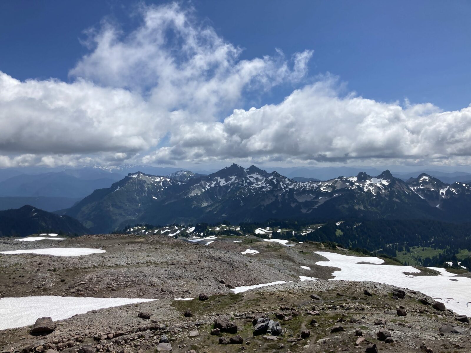 In the foreground, a volcanic landscape of brown rocks and a bit of late season snow along the Skyline Trail. In the background are a range of mountains with some snow still holding on. There are broken clouds against a blue sky.