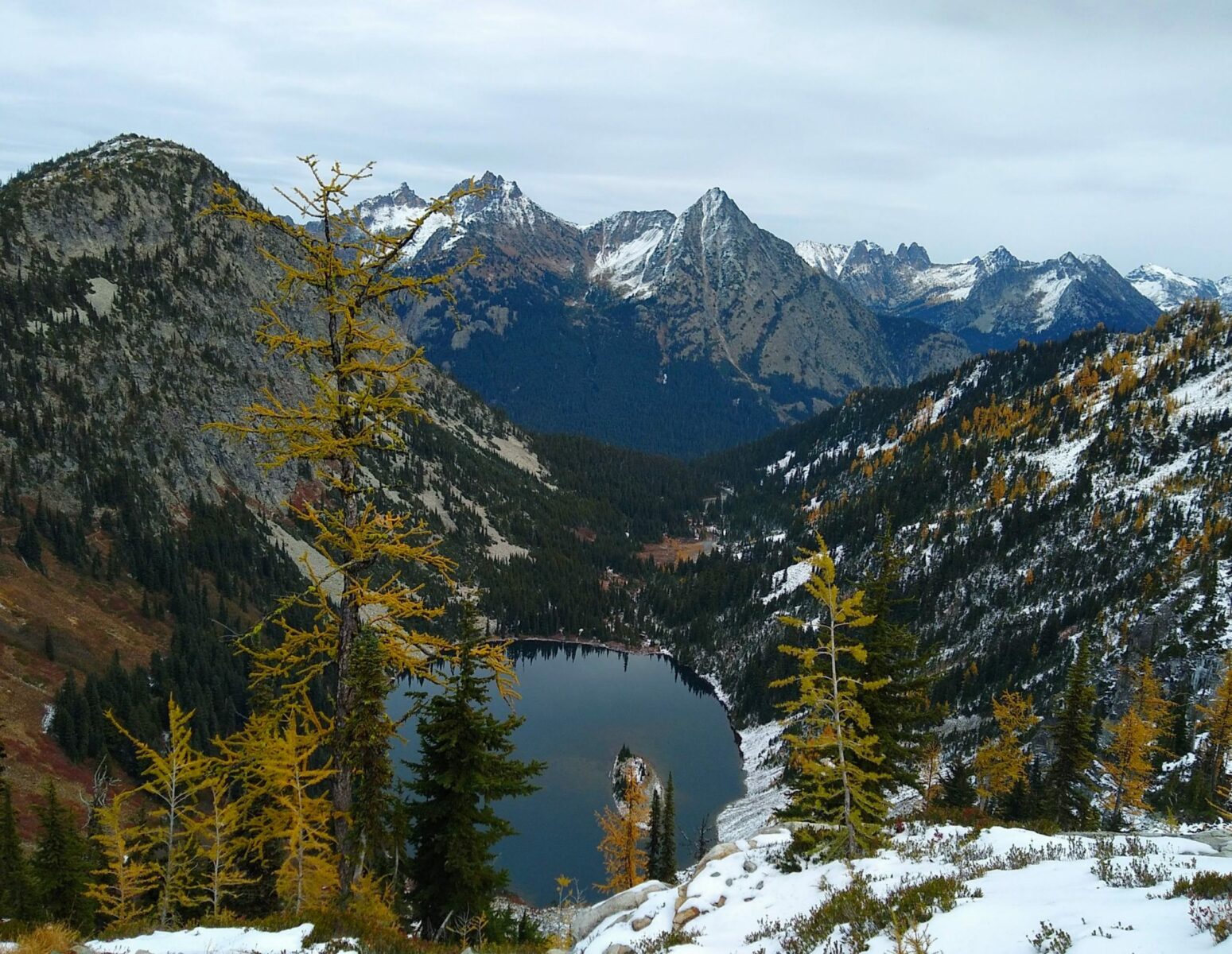 An alpine lake surrounded by forested mountains on the Maple Pass Loop in the North Cascades. There are golden larch trees in the foreground and high snowy mountains in the background