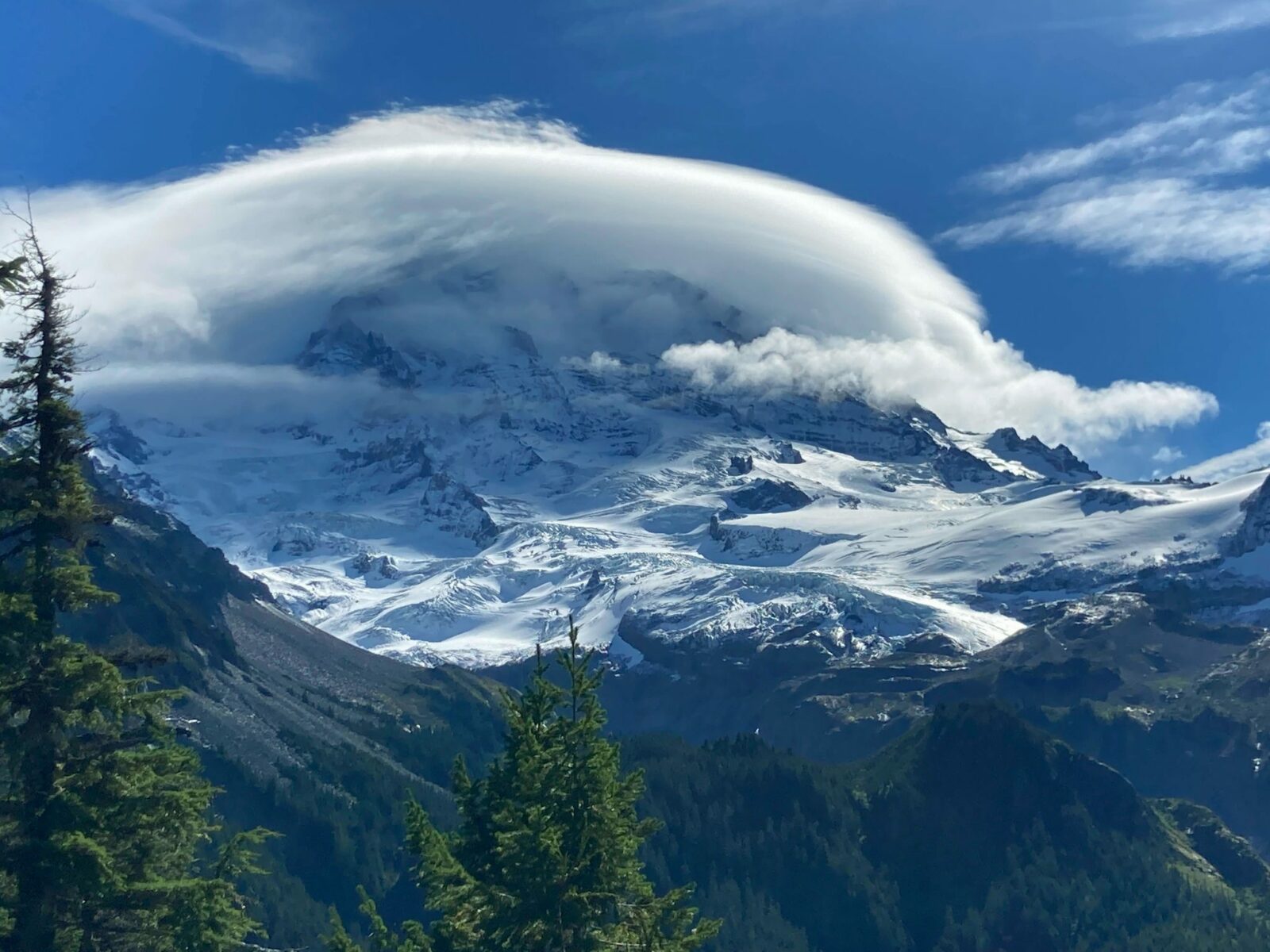 Mt Rainier, a high snowcovered mountain has clouds around it's summit and sides. There are glaciers and rocks visible on the mountain. In the foreground are forested hillsides.