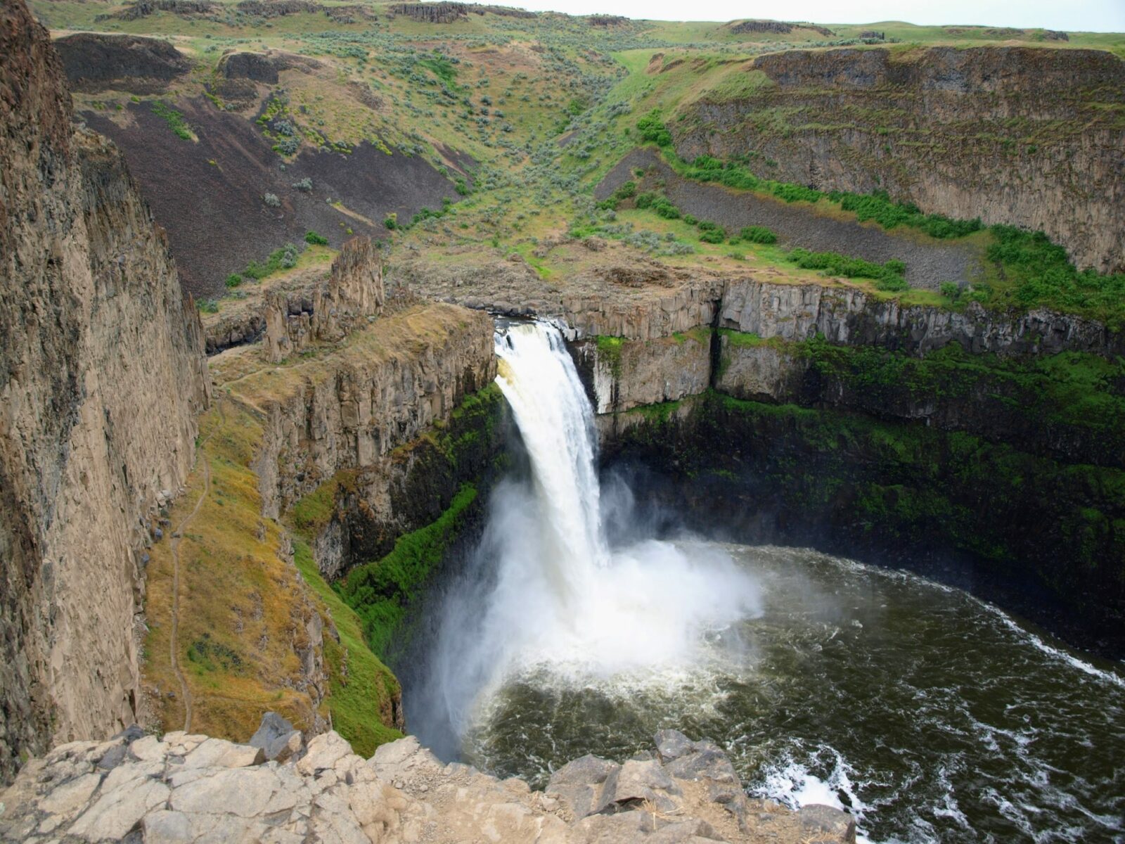 A high waterfall in washington state plunges down a straight rock wall into a pool. There are a few shrubs around on the otherwise dry land above it.