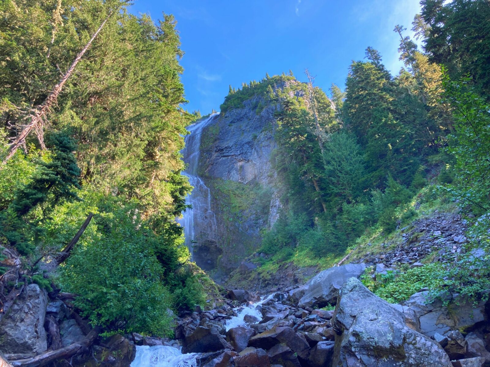 A steep and wide waterfall rolls over a cliff surrounded by forest. There are rocks and a creek in the foreground along the Spray Park trail in Mt Rainier National Park