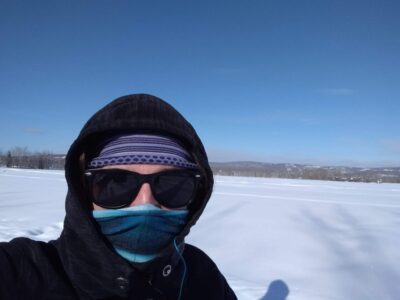a person wearing a fleece jacket, a blue head band, sunglasses and a buff over their nose and mouth in a snowy field on a sunny winter day in Fairbanks Alaska