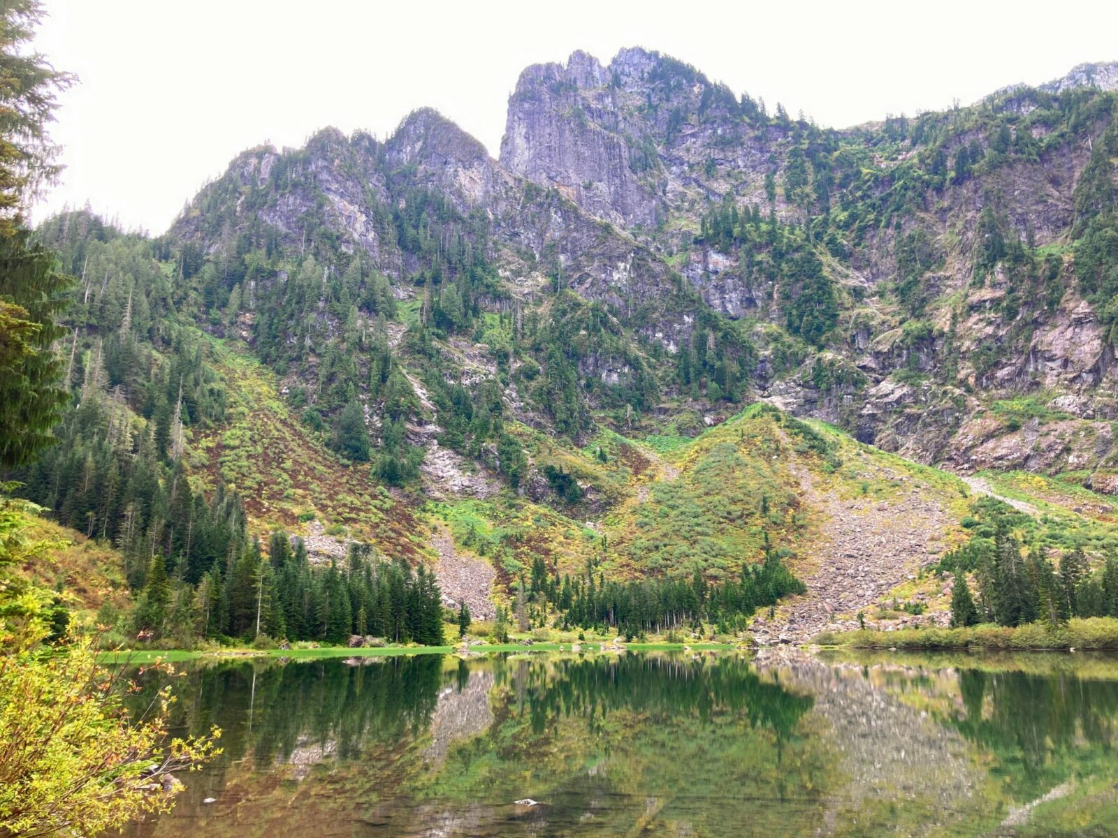 A small glassy alpine lake with trees and mountains reflected in it. The lake is surrounded by high mountains and evergreen trees and rocks