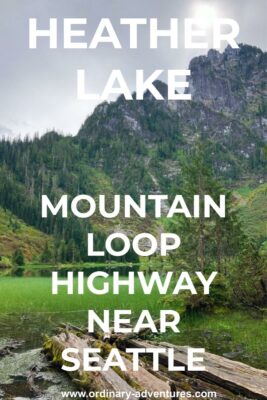 Green grass grows out of Heather Lake, a small alpine lake. There are logs in the foreground and a high mountain in the background. There are also trees and shrubs around the lake, Text reads: Heather Lake Mountain loop highway near Seattle