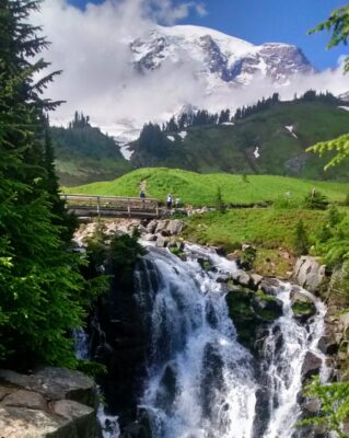 A waterfall with a bridge over it and a few people on a hiking trail. In the background is Mt Rainier, and a few clouds surround it. There are trees and a green meadow beside the trail and waterfall.