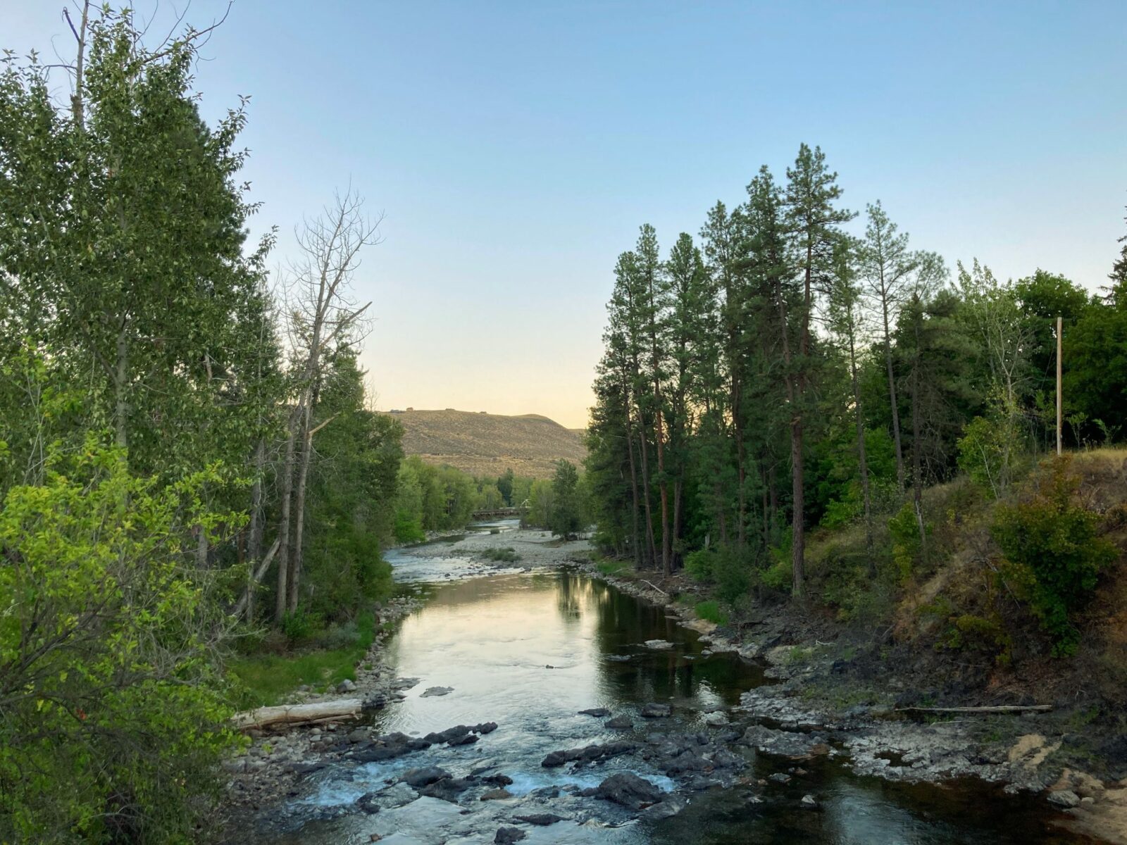 A peaceful river with trees on each side at dusk in the town of Winthrop, Washington. In the distance is a dry brown hill.
