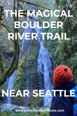 A high waterfall coming down a rock cliff surrounded by moss and ferns. There are evergreen trees in the foreground and a hiker with an orange hat looking at the waterfall. Text reads: The magical Boulder River Trail near Seattle