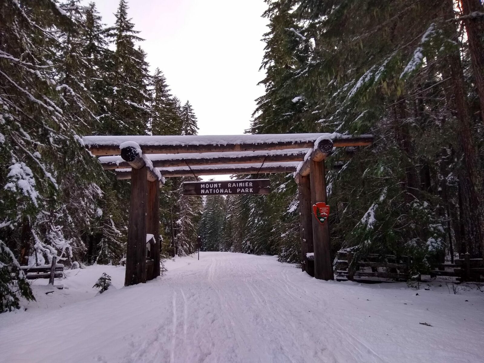 A road covered in snow surrounded by evergreen trees that are also snow covered. A wooden sign over the road says Mount Rainier National Park