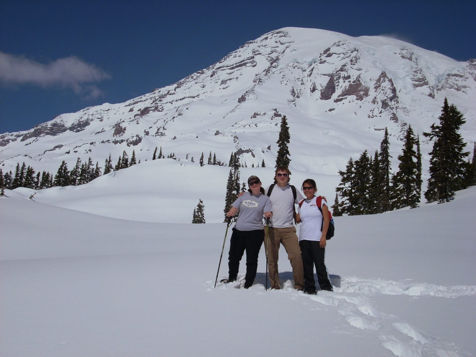 Three hikers pose for the camera in snow in front of Mt Rainier. They are wearing snowshoes, t shirts and sunglasses. Evergreen trees are visible as well.