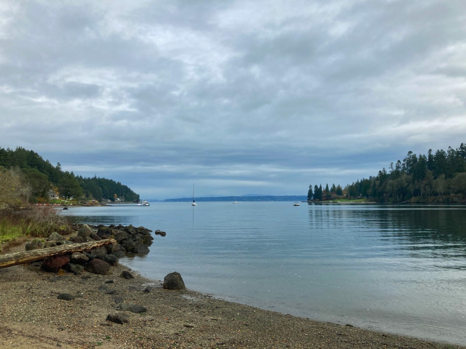 One of the hikes on bainbridge island leads from Fort Ward to the head of Blakeley Harbor in Blakeley Harbor park. The park has a rocky beach that is surrounded by evergreen trees. The harbor is shallow and narrow, with a few boats anchored in the distance.