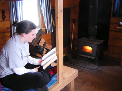 Camping in Alaska in a public use cabin. A person is sitting on a bunk reading a book next to a woodstove burning in the wooden cabin across the room and a window with open curtains to let in the sun