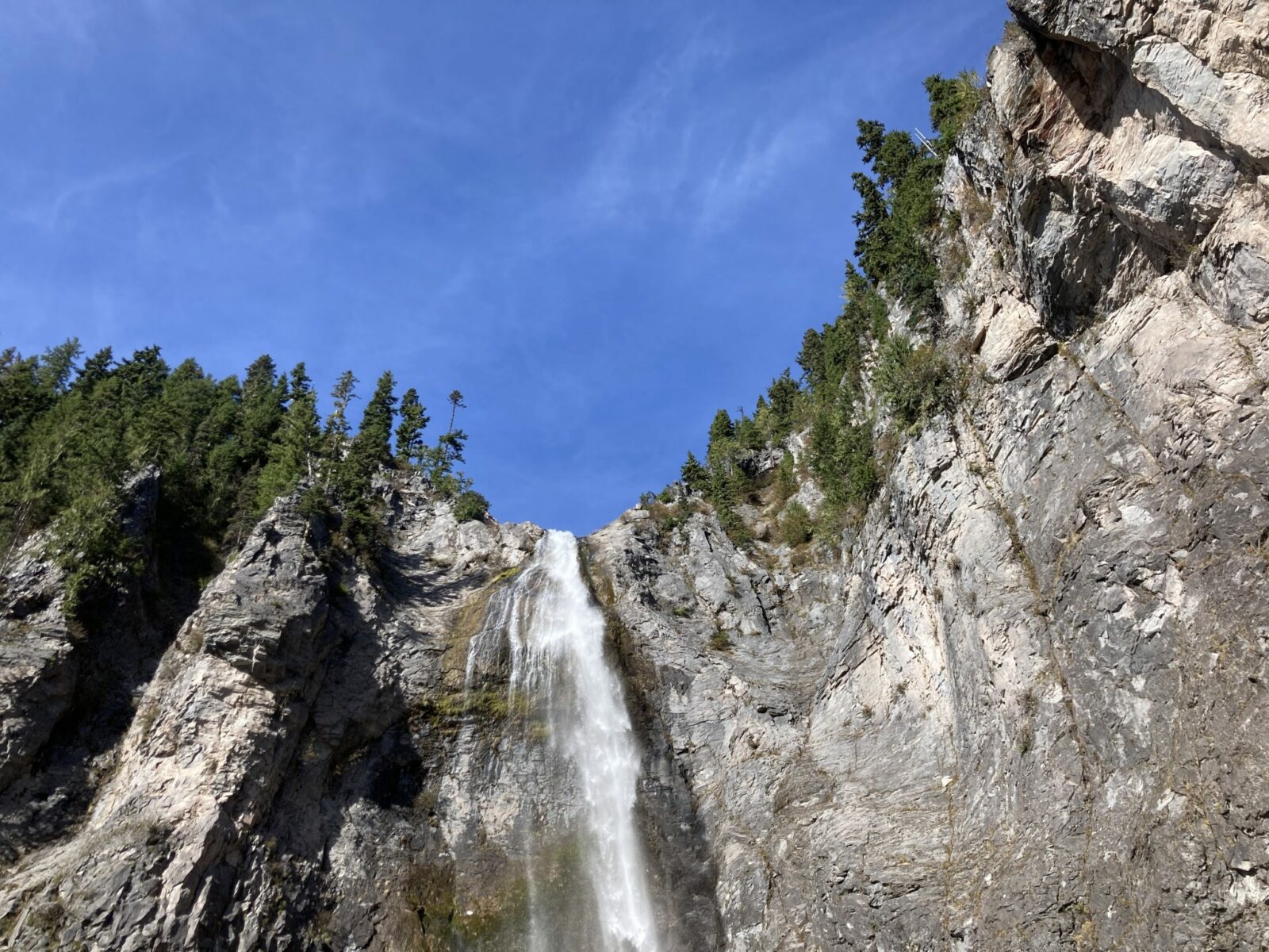 Looking up at a high waterfall coming over a cliff face. There are evergreen trees lining the top of the cliff and the sky is blue