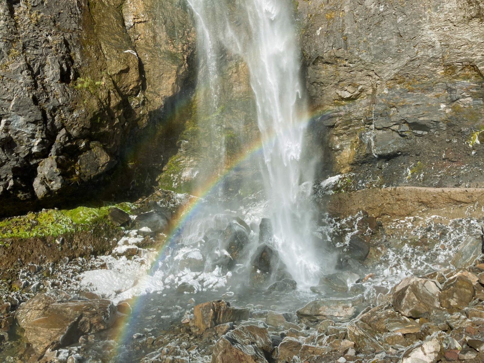 The base of Comet Falls crashes onto frosty rocks. There is also foam from the waterfall and a rainbow arcs across the base of the waterfall
