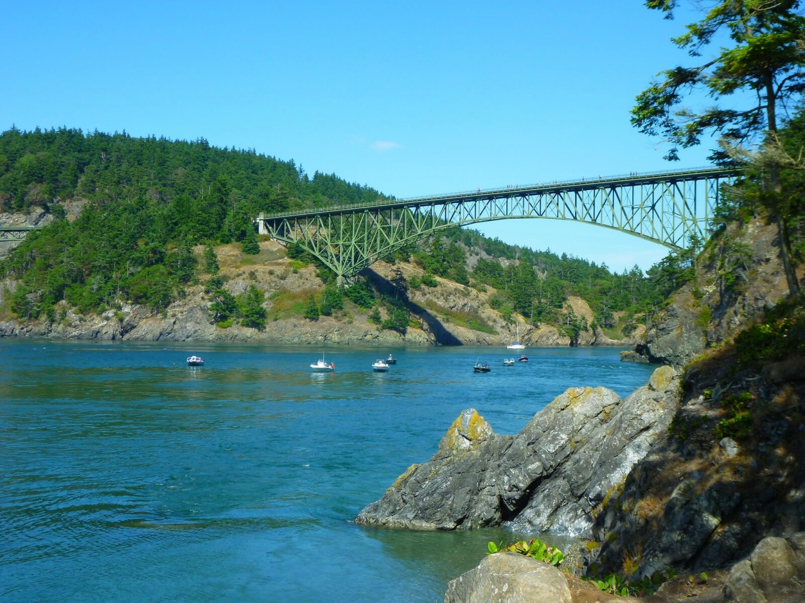 A green steel bridge is high above the water between two rocky and forested hills on each side of a narrow passage. There are a few fishing boats and one sailboat in the water under the bridge