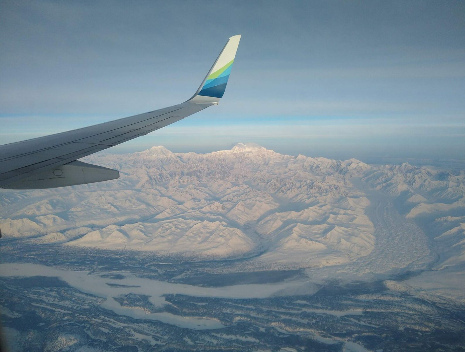 An airplane wing with Denali, glacier, other mountains and the Susitna River below, flying over Alaska in winter