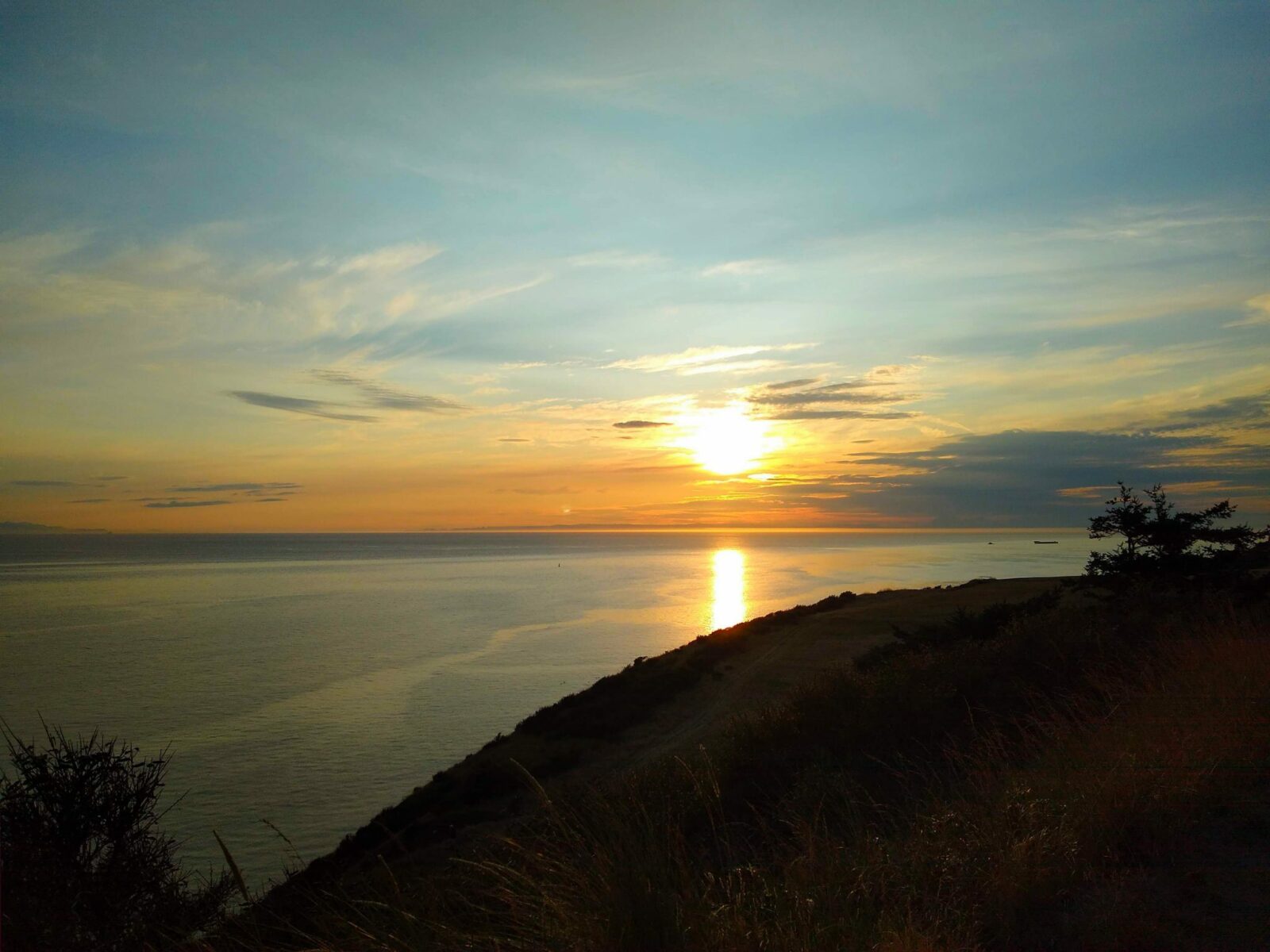Sunset from a whidbey island hike at Fort Ebey State Park. There is a bluff in the foreground in shadow with a couple of trees. Beyond is calm water going as far as you can see. The sun is just setting behind a few clouds