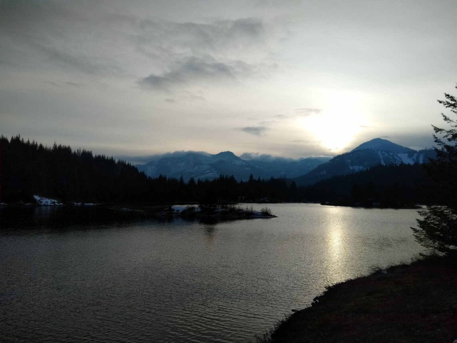 A winter sun sets on a day with high overcast behind a mountain near gold creek pond. The pond is surrounded by forest and forested mountains in the distance
