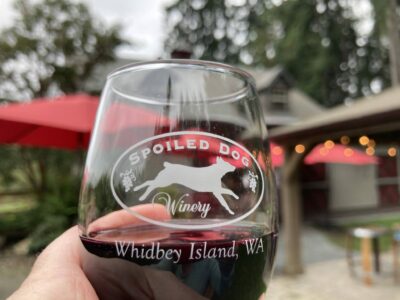 A close up of a glass of red wine. The wine glass has a picture of a dog and says Spoiled dog winery, Whidbey Island WA. In the background are out of focus tables, red umbrellas and outdoor lights.