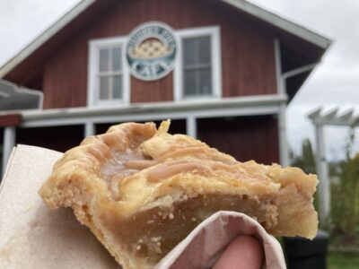 A close up of a piece of apple pie with a crust and caramel on the top. In the background is an out of focus red building