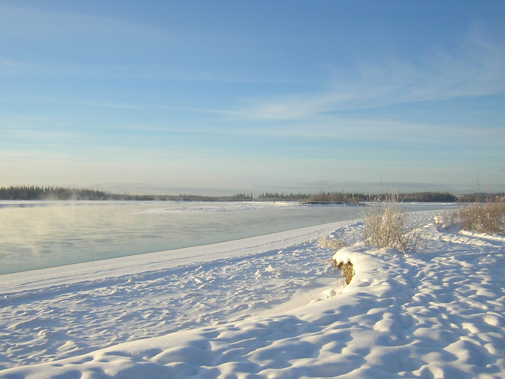 A frozen river in Alaska in winter. There are forests across the river in the distance and a snowy shore in the foreground.