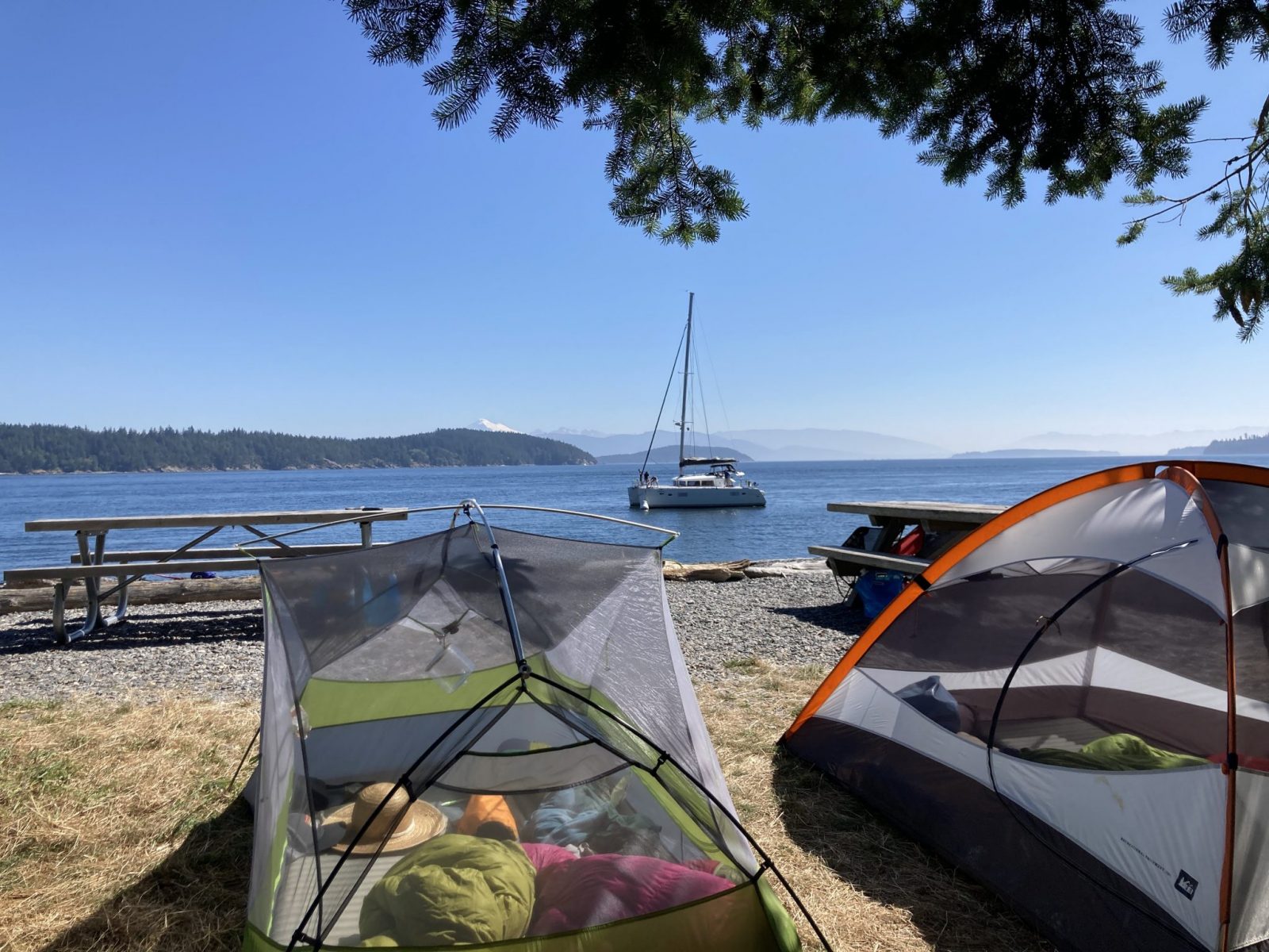 Camping in the San Juan Islands at Pelican Beach. There are two tents with their rain flies off to catch the sun. There are picnic tables and a rocky beach in background. A sailboat is anchored just off shore. In the distance are forested islands and a high snow covered mountain.