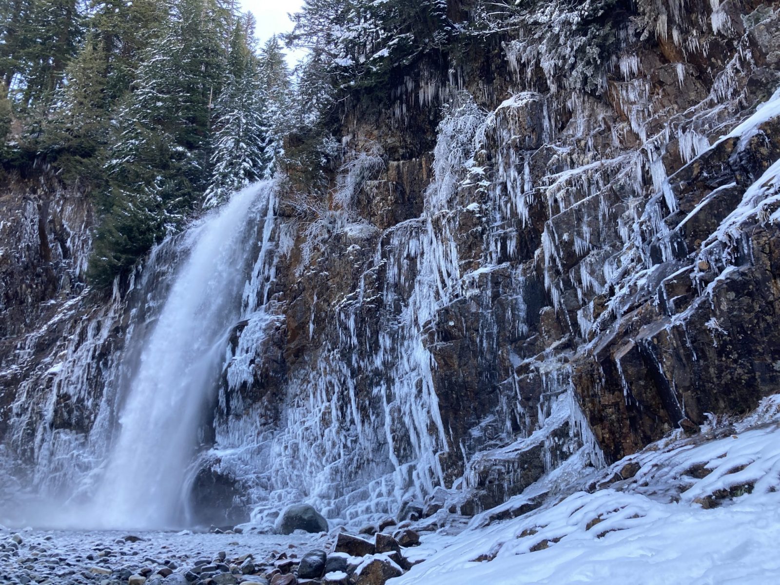 A powerful waterfall coming over a vertical rock face. The rocks are covered in icicles and the ground around the waterfall is covered in snow.