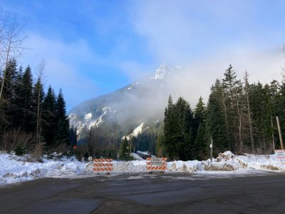 Road closed barricades in front of a snow berm on a closed road. In the distance is a mountain partly covered by fog and there are evergreen trees in the foreground.