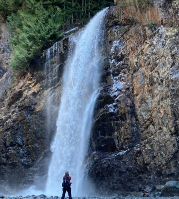 Franklin Falls in winter with only a bit of ice and snow around it. The waterfall is plunging over a vertical cliff. A hiker is taking a photo of the waterfall wearing a wool hat and a red jacket