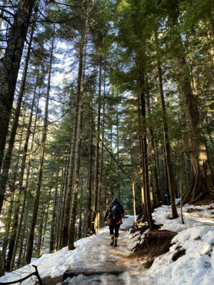 A hiker walking on the trail to Franklin Falls in winter. There is packed ice on the trail and it is surrounded by evergreen trees. The hiker is wearing a blue and green hat, a brown down coat, black leggings, rubber boots and a blue backpack