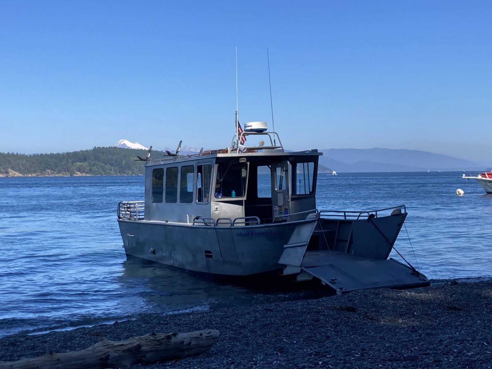 A metal charter boat is beached with it's ramp down to let hikers disembark. It's a gravel beach in the San Juan Islands with forested hillsides and a high mountain in the distance