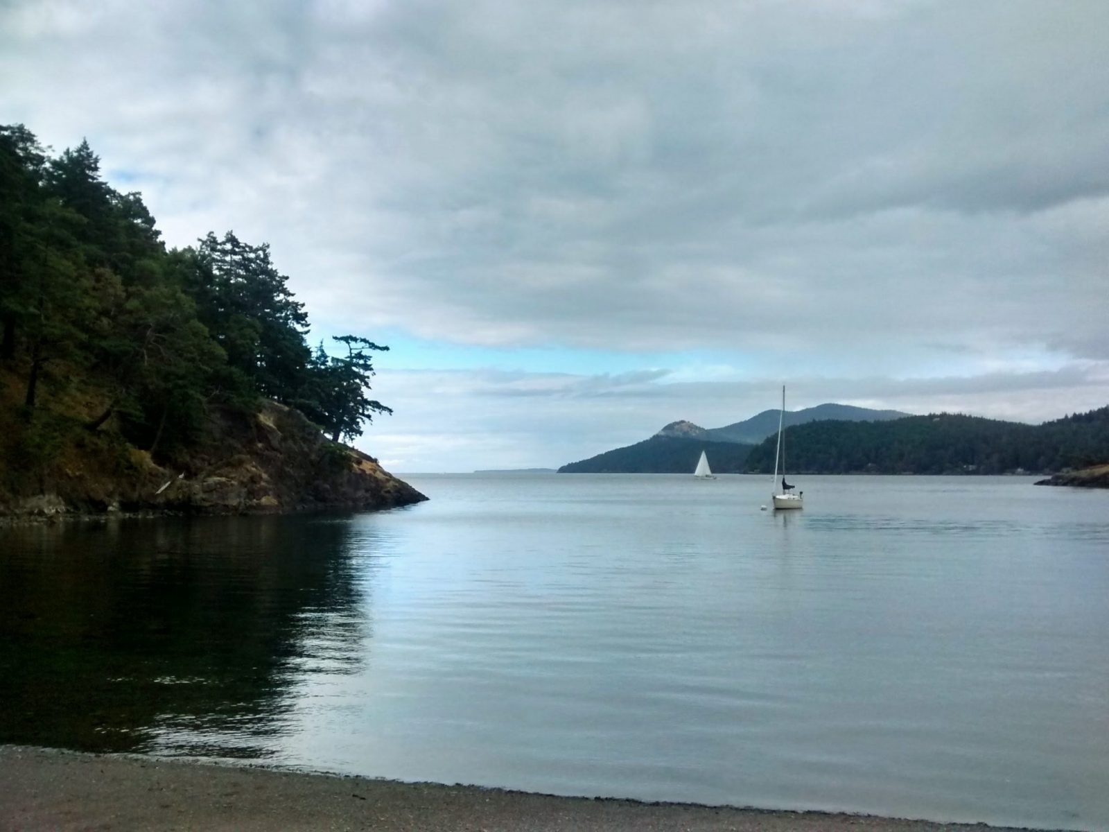 A sailboat at anchor in a small bay. The bay is surrounded by forested hillsides and a gravel beach on a cloudy day