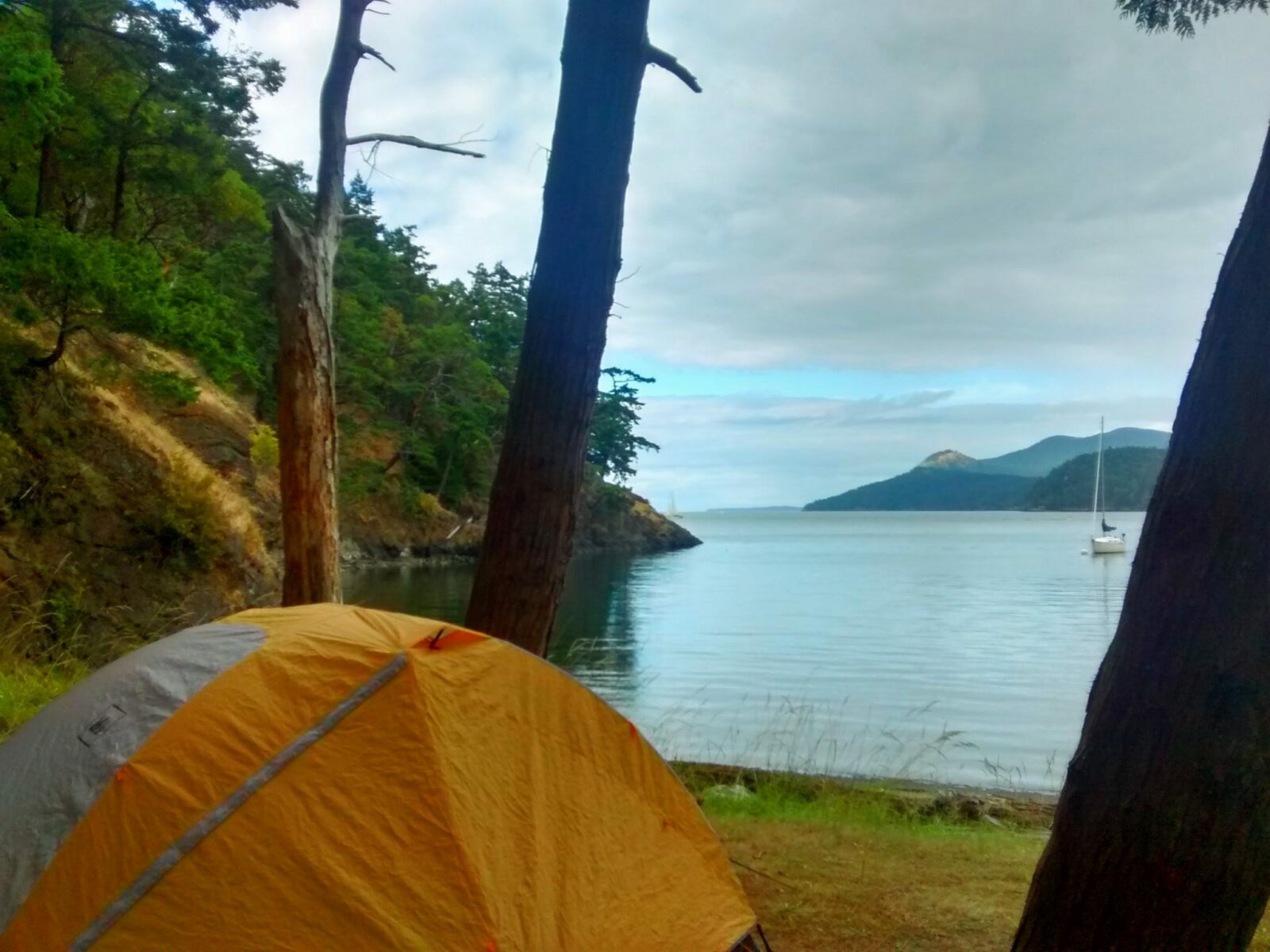 Camping in the San Juan Islands on Jones Island. An orange and white tent is between a few trees near the beach. There are green trees along the shore and a sailboat anchored in the bay. In the distance are more forested islands