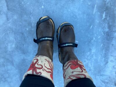 A person's brown rubber boots with an octopus print around the cuff with microspikes attached to the boots. The boots are standing on ice.