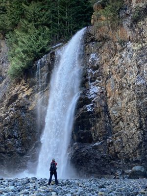 A photographer stands in front of Franklin Falls in winter. At the base of the falls are icy rocks and the waterfall plunges over a vertical cliff with evergreen trees at the top. There are a few small icicles around the waterfall