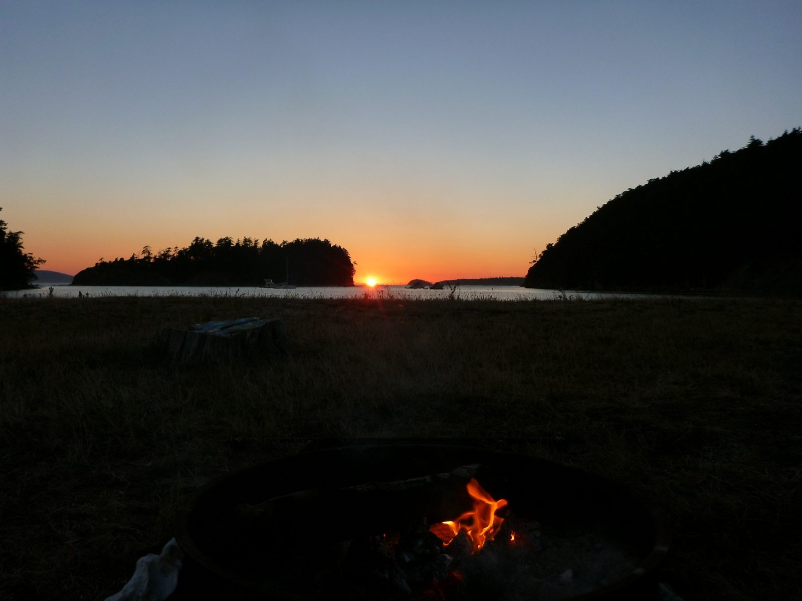 A sunset between distant islands and a campfire in the foreground, camping in the san juan islands