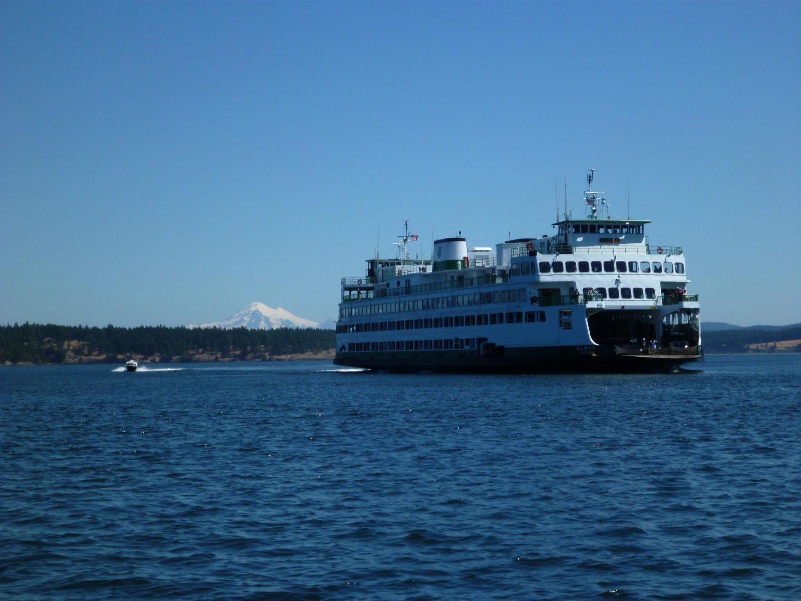 A white Washington State ferry with an open front end showing cars with a small powerboat next to it. There is a forested island in the background and a high snow capped mountain