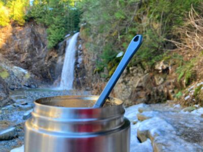 A close up of a thermos with hot food and a spoon. Franklin Falls is in the background. There is ice and snow around on the trail.