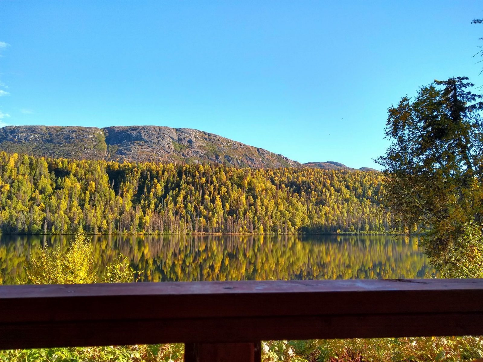 A wooden railing in the foreground with a calm lake surronded by trees and hills