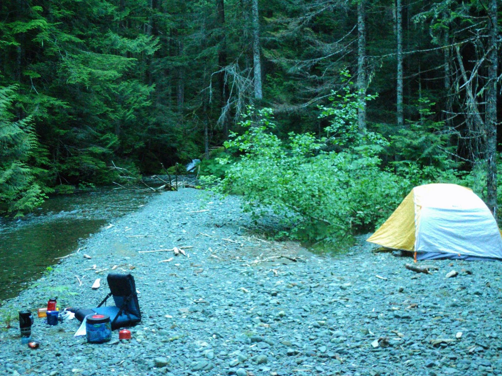 A gravel bar next to a creek in an evergreen tree forest. There is a yellow and white tent set up on the gravel and camping equipment for cooking dinner nearby.