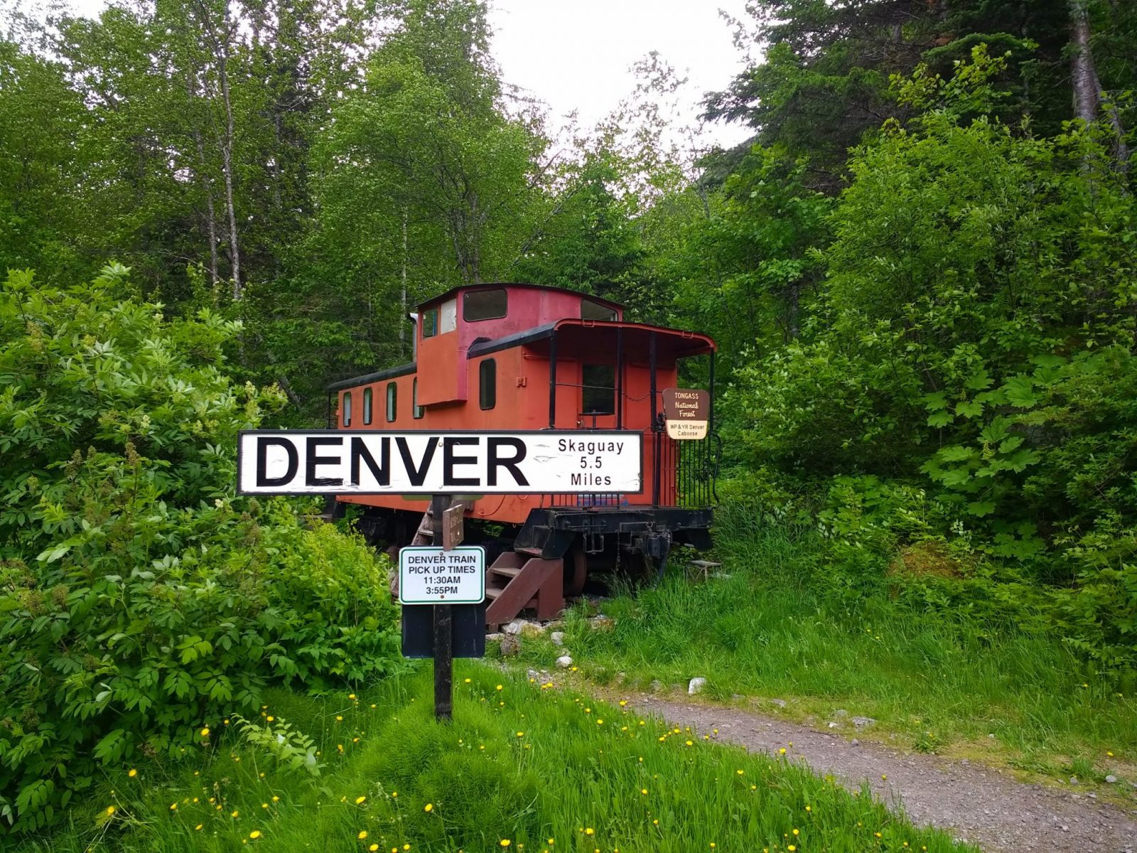A red train caboose next to a trail. A railroad sign says "Denver" in the foreground. The sign and caboose are surrounded by green forest. For a unique camping experience in Alaska you can sleep in the caboose