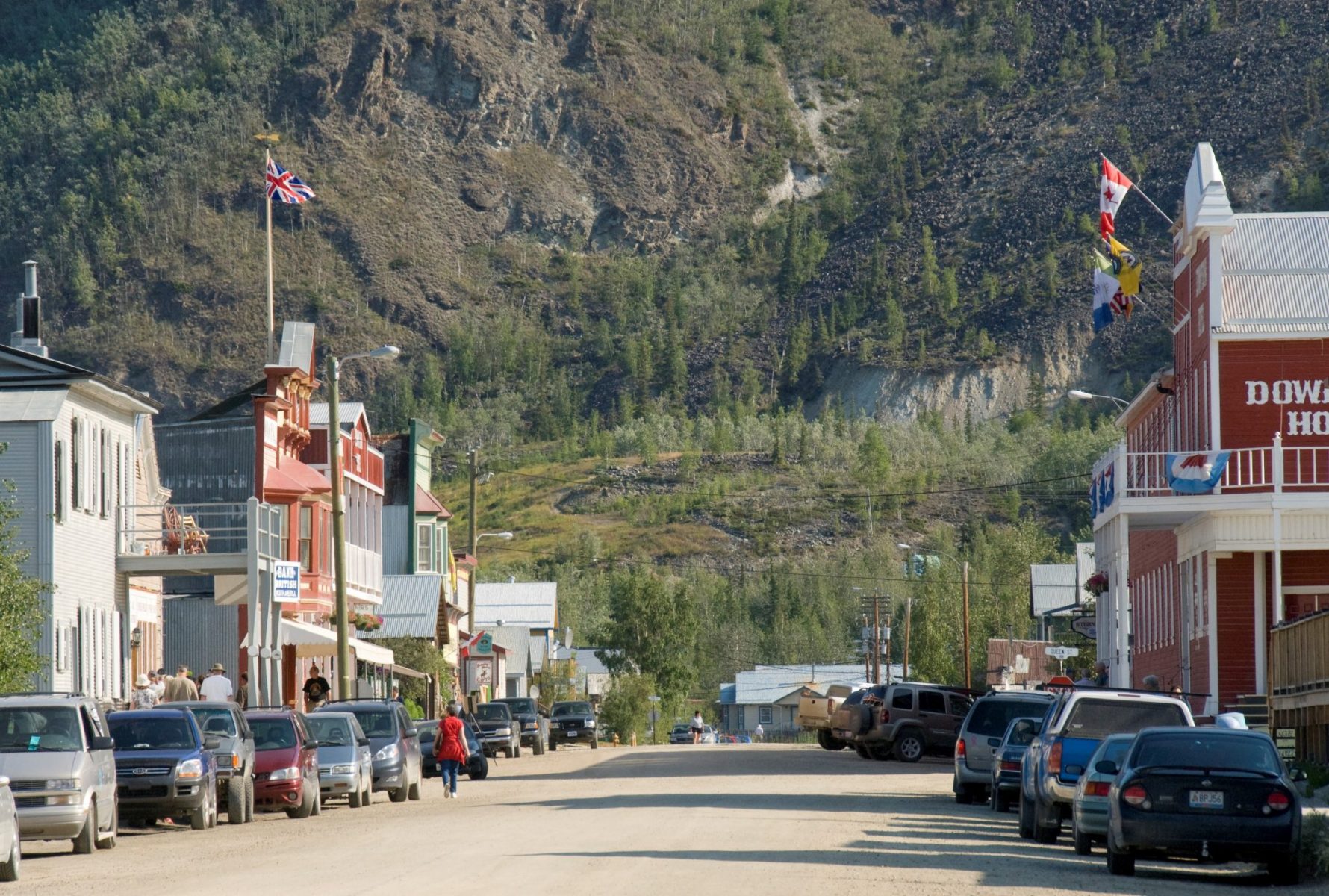 A gravel street in a small town lined with parked cars. The buildings are restored historic buildings. In the background in a forested hillside. Walking the historic streets is one of the best things to do in Dawson City