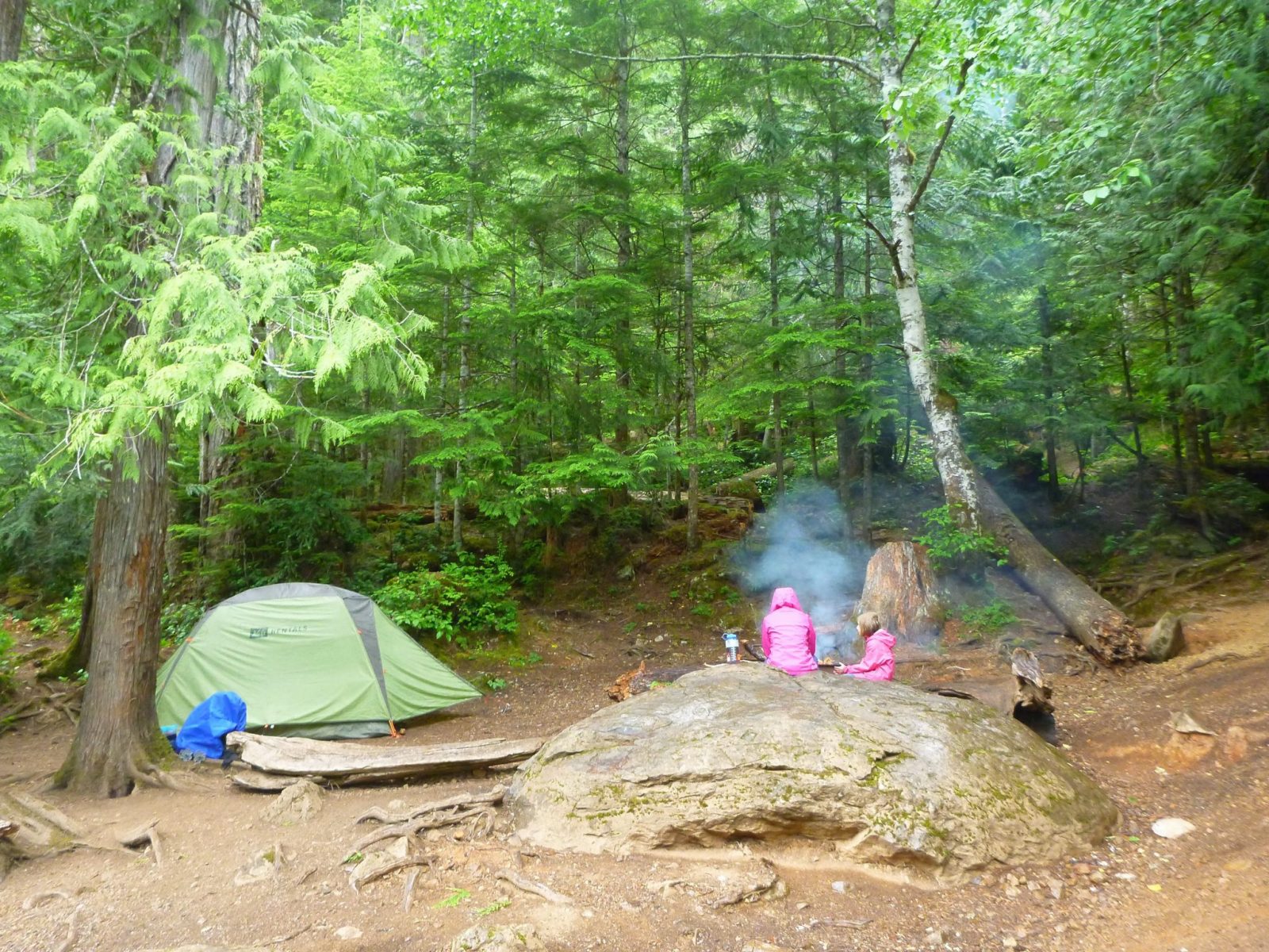 An adult and a child wearing pink rain coats sitting next to a campfire. There is a big boulder next to them and a green tent. The campsite is surrounded by trees.