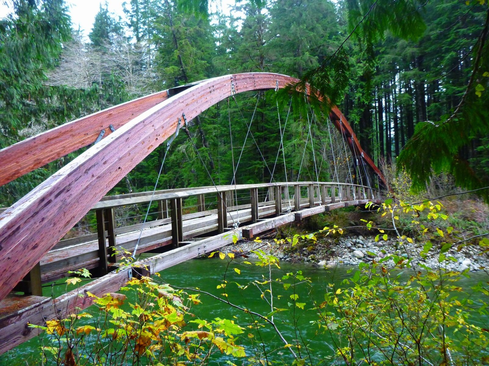 A wooden and cable bridge over the middle fork snoqualmie river, one of the many hikes near north bend washington, with an arched wooden top over a fast moving river. The bridge is surrounded by evergreen trees and bushes.