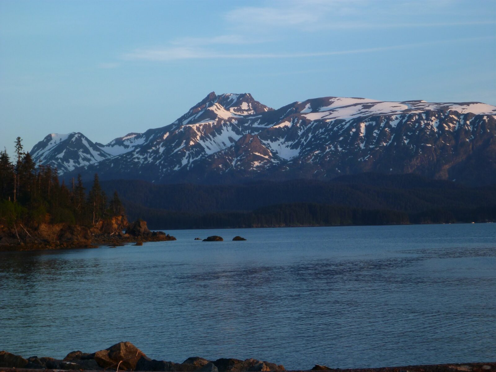 Evening sun hits mountains with lingering snow across a bay. In the foreground are trees and a rocky shore