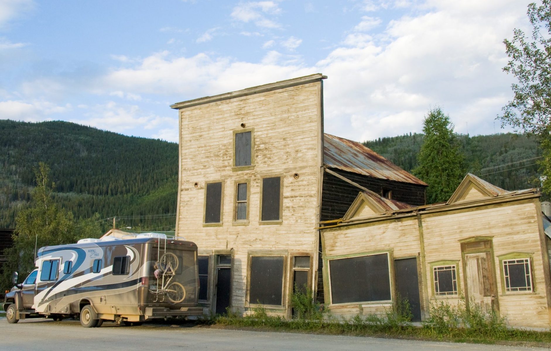 Looking at old buildings is one of the best things to do in Dawson City, here three brown buildings from the late 1890s are boarded up and leaning over slightly. In front of the buildings is a motorhome covered in mud