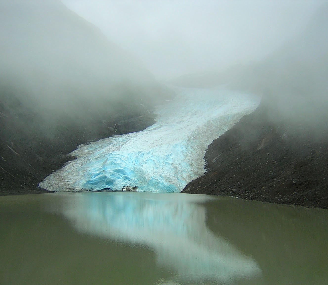 The toe of a glacier at the edge of a lake. The glacier is between two sides of a rocky hillside. It's a foggy day