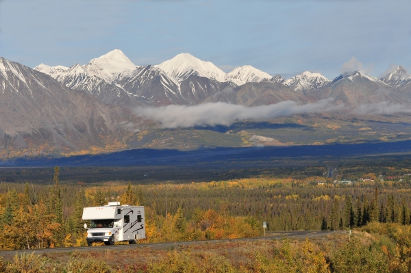 The snowcapped St Elias mountain range and forests with some fall color surround an RV on a two lane highway on the drive to alaska